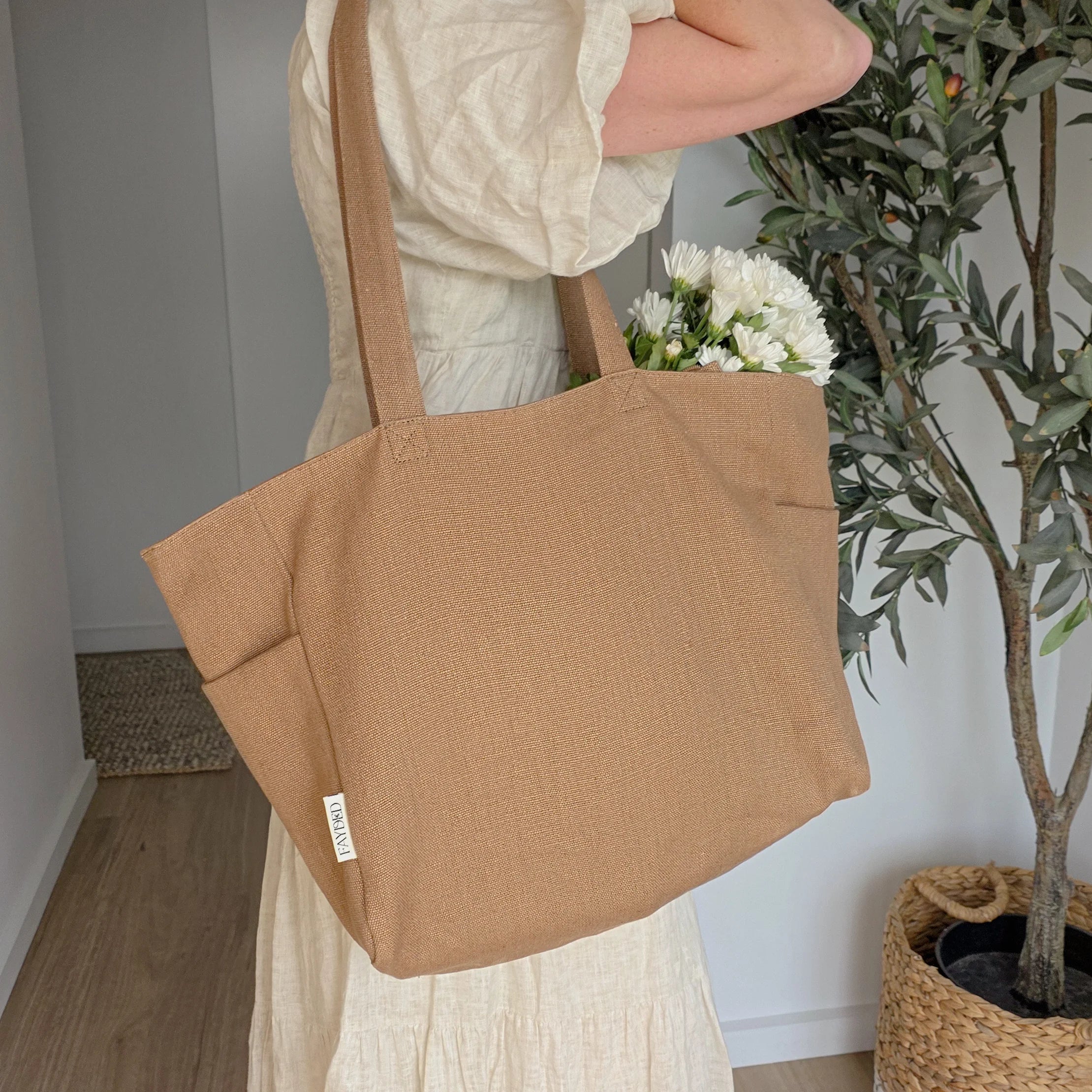 Person holding a brown tote bag with flowers, standing indoors next to a plant.