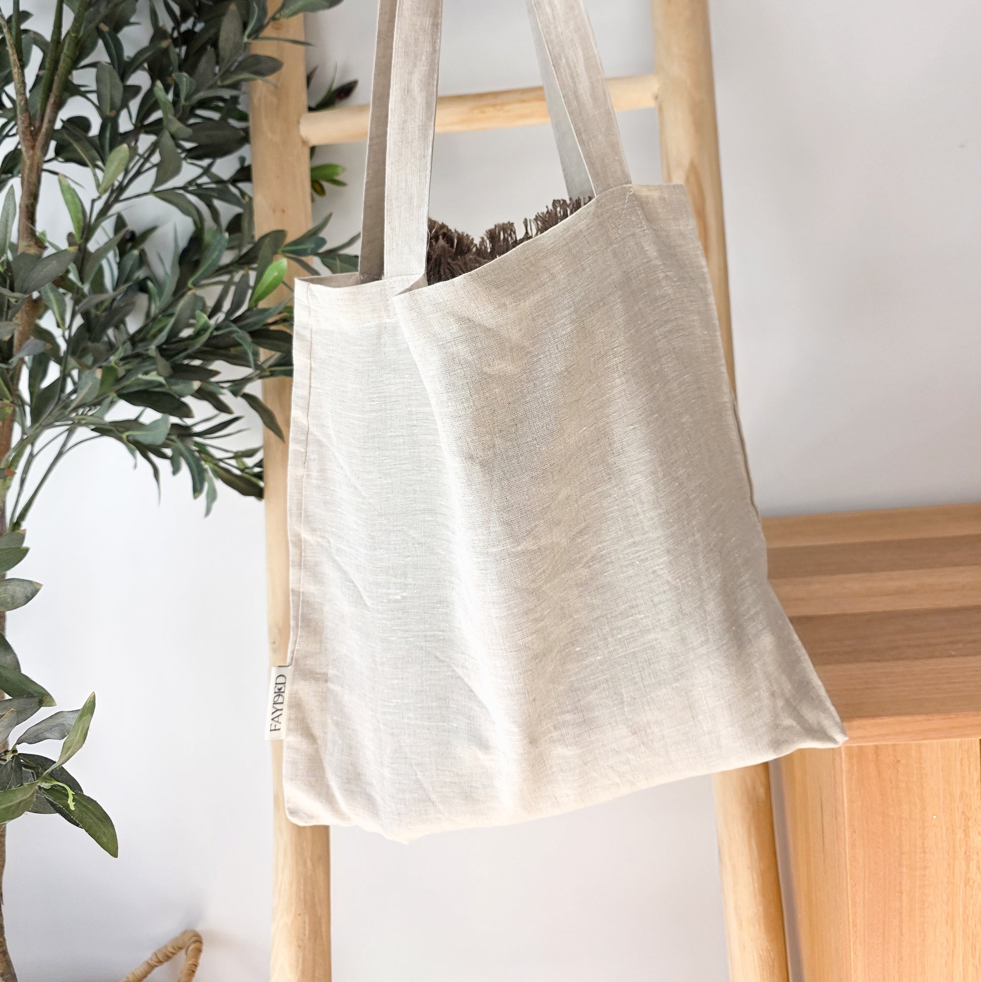 Beige tote bag hanging on a wooden ladder with a plant in the background