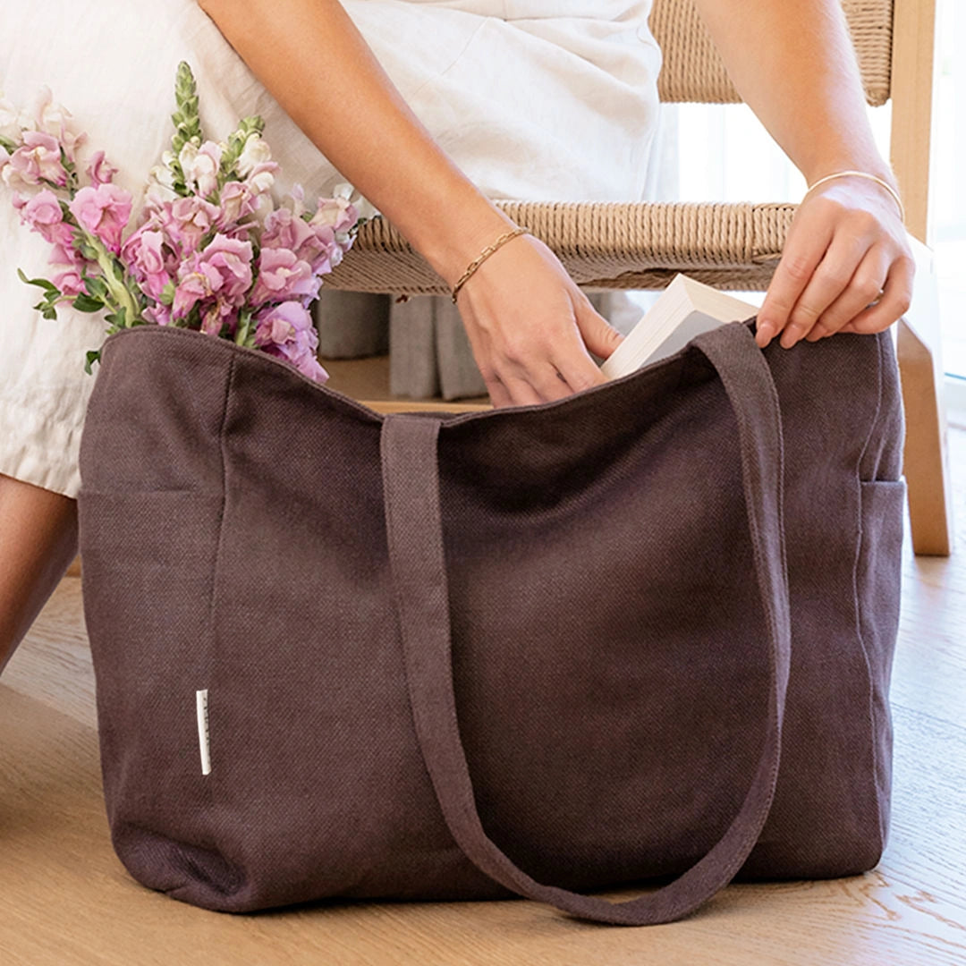 Person opening a brown tote bag with flowers and books inside on a wooden floor.