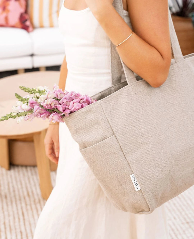 Person holding a beige tote bag with flowers, wearing a white top and beige pants. 100% linen. Natural fibre bag