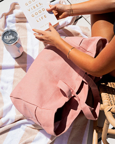 Person holding a pink tote with a towel and tumbler nearby a pool setting