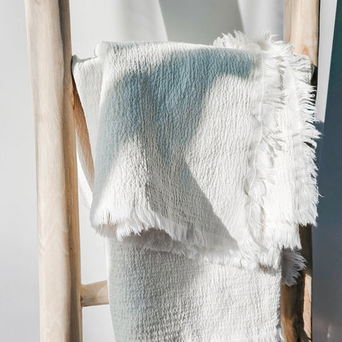 Textured blanket with fringe draped over a wooden ladder against a white background